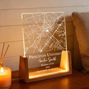 以下が含まれることがあります： プリンストン大学の地図が刻まれた照明付きの装飾品。Amelia Smith、Bachelor of Art 2025、座標が記載されています。透明なアクリルパネルに地図がエッチングされ、木製の台座に設置されています。
