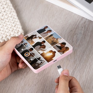 Puede incluir: Un marco de fotos digital rosa que muestra un collage de fotos de boda. El marco tiene forma cuadrada con esquinas redondeadas y un puerto USB-C. Un cable USB-C blanco est&aacute; enchufado. La pantalla muestra una interfaz de reproductor de m&uacute;sica.