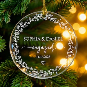May include: Clear, round Christmas ornament with a gold string, featuring the names "Sophia & Daniel" and the word "engaged" in elegant script. The date "11.16.2025" and a heart are also included, surrounded by a white floral wreath.