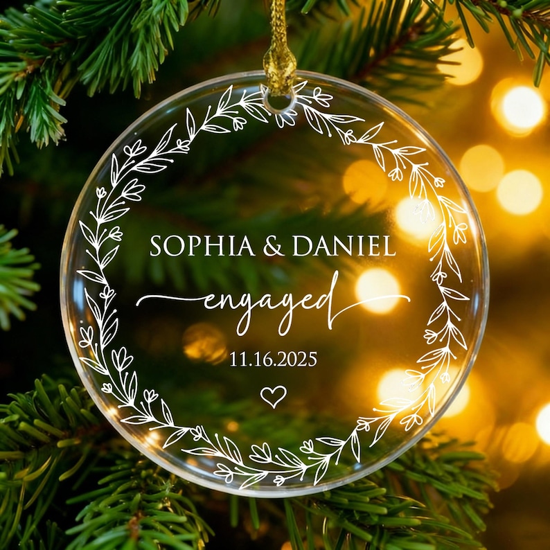 May include: Clear, round Christmas ornament with a gold string, featuring the names "Sophia & Daniel" and the word "engaged" in elegant script. The date "11.16.2025" and a heart are also included, surrounded by a white floral wreath.