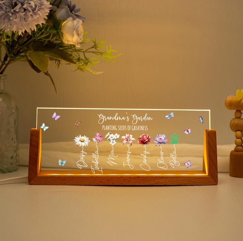 May include: A wooden-framed LED light with the text "Grandma's Garden" and the phrase "Planting seeds of greatness." The clear panel features colorful flowers, butterflies, and names. The base is a warm, natural wood tone.