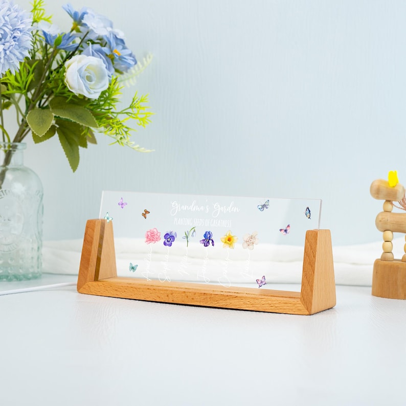 May include: A clear acrylic sign with the text "Grandma's Garden" and colorful flower illustrations, supported by a light-colored wooden base. Butterflies are scattered around the flowers. A vase of blue flowers is in the background.