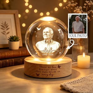 May include: A spherical glass ornament featuring an engraved portrait of an older man, the word "dad," and the dates "1946-2026." The glass sphere sits on a circular wooden base engraved with the text "Those we love don't go away they walk beside us every day."