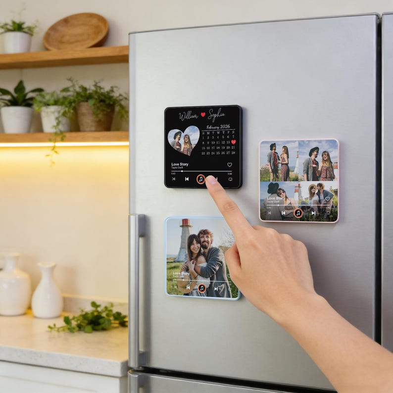 Puede incluir: Un dedo toca un im&aacute;n cuadrado negro con una foto, un calendario y una interfaz de reproductor de m&uacute;sica. El im&aacute;n est&aacute; en la puerta plateada de un refrigerador, con otros dos imanes con fotos. El im&aacute;n superior tiene los nombres "William & Sophia" y el texto "Love Story".