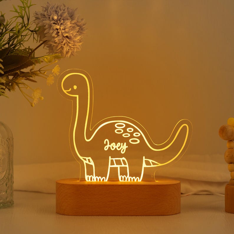 May include: A glowing acrylic dinosaur night light with a warm yellow illumination. The dinosaur silhouette features the name Joey in script lettering on its side, mounted on a smooth, oval-shaped light-coloured wooden base.