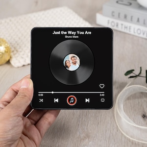 Puede incluir: Placa musical cuadrada negra con la canci&oacute;n "Just the Way You Are" de Bruno Mars. El dise&ntilde;o incluye una imagen circular de una pareja, un bot&oacute;n de reproducci&oacute;n y una barra de progreso. La placa se sostiene en una mano.