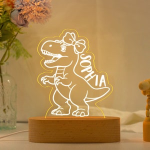 May include: A white dinosaur night light with a pink bow on its head. The dinosaur is outlined in yellow and the name "Sophia" is written on its side. The light is on a wooden base.
