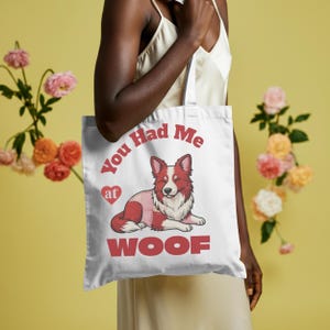 May include: White tote bag with a red and white cartoon dog and the text "You Had Me at WOOF." The dog has a heart next to the word "at." The bag has white handles. The bag is being held up.