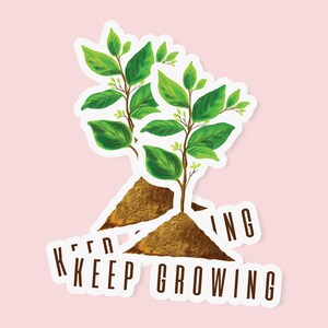 May include: Two stickers featuring a plant with green leaves and brown soil. The text "KEEP GROWING" is printed in brown on a white background. The stickers are on a light pink background.