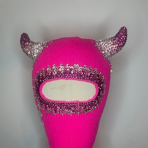 Könnte beinhalten: Eine rosafarbene Strickbalaclava mit silbernen und rosafarbenen Strasssteinen, mit zwei Hörnern oben.