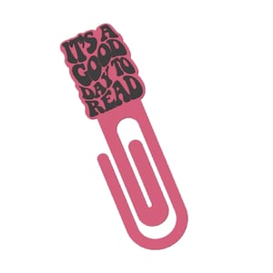 May include: A pink metal bookmark shaped like a paperclip, featuring the text "IT'S A GOOD DAY TO READ" in black. The bookmark has a rectangular top section. The text is in a retro, bubbly font.