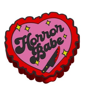Può includere: Una spilla a forma di cuore con un bordo smerlato rosso e le parole "Horror Babe" in corsivo nero su sfondo rosa. Un piccolo coltello con una lama rossa è presente sotto il testo, con accenti di stelle gialle.