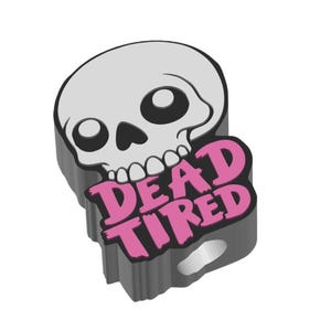 May include: A gray skull-shaped charm with the words "DEAD TIRED" in pink. The charm has a black border and a hole for attaching to a keychain or other item. The skull has large, round eye sockets and a row of teeth.
