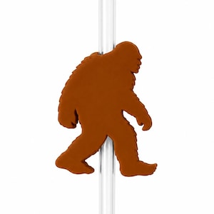May include: A clear, cylindrical drinking straw featuring a brown, flat, silicone-style attachment shaped like a silhouette of a Bigfoot or Sasquatch figure. The creature is depicted in a walking profile, centered on the vertical straw.