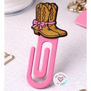 May include: A pink paperclip with a decorative cowboy boot design. The boots are tan with brown trim and a pink bow. The paperclip has the text "MFL STL FILES EST. 2024" in a circle.