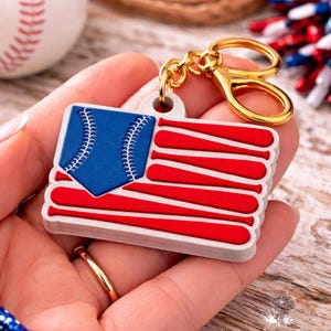 Puede incluir: Un llavero con temática de béisbol que presenta un diseño que combina una pelota de béisbol y bates para parecerse a la bandera estadounidense. El llavero tiene un cierre y una cadena dorados. La parte de la pelota de béisbol es azul con costuras blancas.