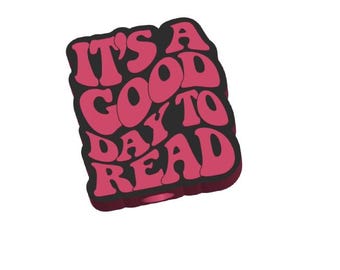 May include: A rectangular sticker with the phrase "IT'S A GOOD DAY TO READ" in a retro font. The text is in a bubble-style font in shades of pink, outlined in black. The sticker is on a white background.