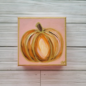 May include: A small square canvas painting featuring a pumpkin. The pumpkin is painted in shades of orange, brown, and white, with a green stem. The background is a soft pink, and the edges are trimmed with gold.