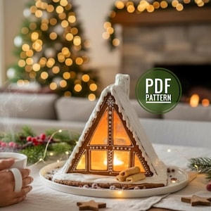 May include: A gingerbread house shaped like an A-frame cabin with white icing details, illuminated from within, resting on a white plate. A green circular badge with the text PDF PATTERN is overlaid on the right side of the image.