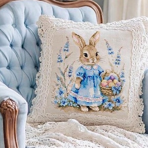 May include: A decorative pillow with a charming bunny design. The pillow features a bunny wearing a blue dress and holding a basket of Easter eggs. The pillow is trimmed with lace and rests on a blue tufted chair.
