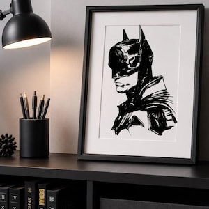 Puede incluir: Una ilustración enmarcada en blanco y negro de Batman de perfil, con una capucha y capa definidas. La obra se presenta en un marco negro fino con un paspartú blanco, situada junto a una lámpara de escritorio negra y varios libros de tapa dura.
