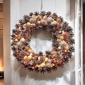 May include: A brown and beige wreath made of pine cones, nuts, and other natural elements. The wreath is hanging on a white door.