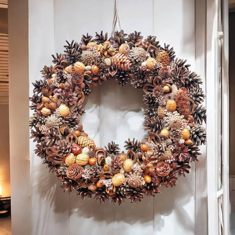 May include: A brown and beige wreath made of pine cones, nuts, and other natural elements. The wreath is hanging on a white door.