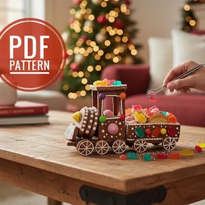 Puede incluir: Un tren de jengibre decorado con caramelos de colores está sobre una mesa de madera. El tren tiene una base marrón con detalles de glaseado blanco y está lleno de varios caramelos. La imagen incluye el texto "PDF PATTERN" en un círculo rojo.