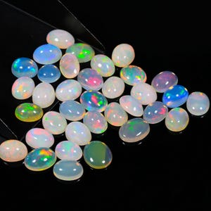 Puede incluir: Una colección de piedras preciosas de ópalo de forma ovalada en varios tonos de blanco, azul y verde. Los ópalos son iridiscentes y tienen un brillo nacarado.