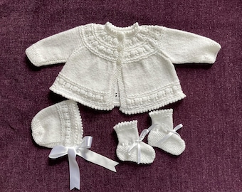 Könnte beinhalten: Ein weißer gestrickter Baby-Cardigan mit Knöpfen, eine passende weiße gestrickte Mütze und zwei Paar weiße gestrickte Booties.