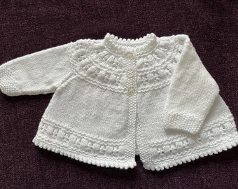 Könnte beinhalten: Weißer gestrickter Baby-Cardigan mit Knopfleiste und einem zarten Spitzenmuster am Ausschnitt und Saum.