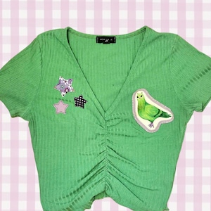 May include: A bright green ribbed short-sleeve top with a V-neckline and a ruched centre front. The garment features three small star-shaped patches in purple and black, and a larger patch depicting a green bird on the right side of the chest.