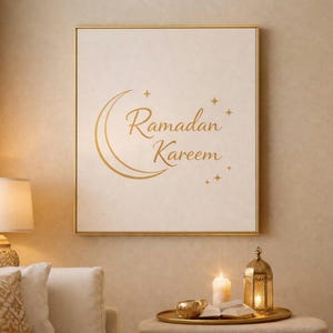 May include: A gold-framed print displaying "Ramadan Kareem" in gold script, with a crescent moon and stars. The print is above a table with a candle, a lantern, and a book. The room has a warm, inviting atmosphere.