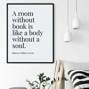 Peut inclure: Impression encadrée en noir avec la citation "A room without book is like a body without a soul." de Marcus Tullius Cicero. L'impression est affichée sur un mur blanc, avec deux lampes suspendues blanches et un coussin à rayures noires et blanches.