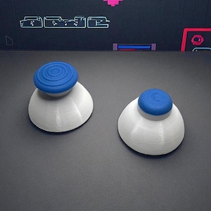 May include: Two white and blue joystick thumbsticks are displayed on a dark surface. The thumbsticks have a white base and a blue, circular top. The background features neon-style graphics.