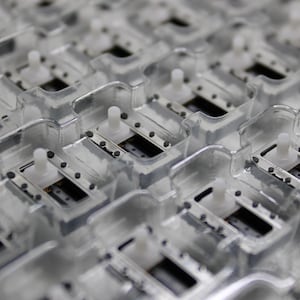 May include: A close-up view of a tray of small, rectangular electronic components. The components are arranged in rows and columns, and each component is encased in a clear plastic housing. The components have a silver-colored metal base and a white plastic top.