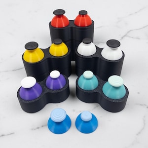 May include: A collection of colorful, 3D-printed bottle stoppers in various colors, including red, yellow, white, purple, teal, and blue. Each stopper has a rounded top and a black base, designed to fit snugly into a bottle opening. The stoppers are arranged on a white marble surface.