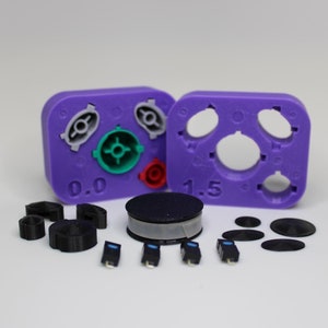 Peut inclure: Des moules en plastique violet avec des cercles de différentes tailles et un interrupteur rond noir avec un anneau blanc. Il y a aussi plusieurs pièces en plastique noir et quatre petits interrupteurs bleus.