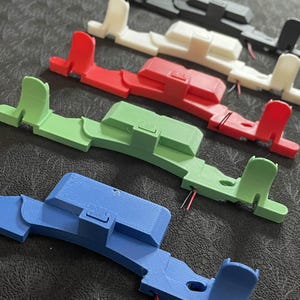 May include: Five 3D printed plastic parts in different colours: black, white, red, green, and blue. Each part has a similar design with a raised section and a small tab on the side.