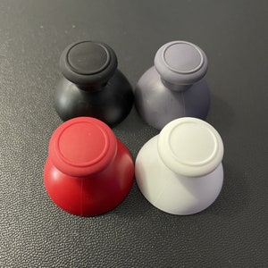May include: Four different colored replacement thumbsticks for a video game controller. The thumbsticks are black, red, white, and gray.