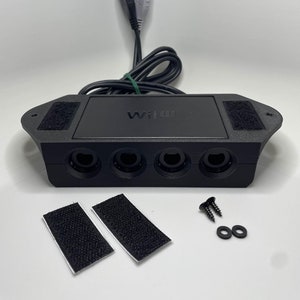 May include: A black Wii U controller adapter featuring four front-facing ports, a top-mounted logo, and an attached black USB cable. The unit includes two rectangular adhesive hook-and-loop strips, two small black screws, and two black washers.