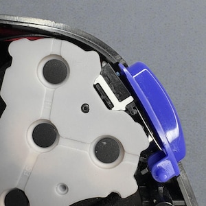 May include: Close-up of a disassembled electronic device component. The image shows a white plastic piece with circular holes, a blue plastic button, and various black and gray internal parts. The device appears to be a controller or similar gadget.