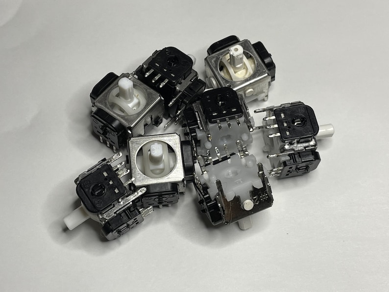 May include: A group of six black and silver analog joystick potentiometers with white plastic shafts. The potentiometers are used in video game controllers.