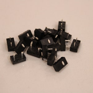 May include: A collection of small, black electronic components with silver pins and white accents. These rectangular components are scattered on a light-colored surface, suggesting a supply of electrical parts.