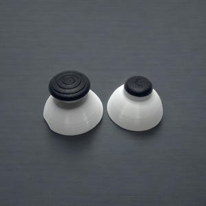 May include: Two white and black bottle stoppers. Each stopper has a white, conical base and a black, circular top. The stoppers are displayed on a gray, textured surface.