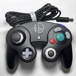 May include: A black GameCube controller with a white Super Smash Bros. logo and white buttons. The controller has a black and white braided cable.
