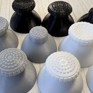 May include: A collection of nine replacement analogue joystick thumbsticks for gaming controllers, arranged in a grid. The thumbsticks are available in black, white, and grey, each featuring a textured, circular top surface with a ridged edge.