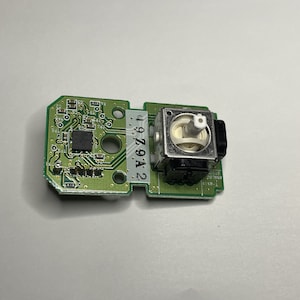 May include: A green circuit board with a small black chip and a silver and black analog stick. The board has the number "2 979A 2 1" printed on it.
