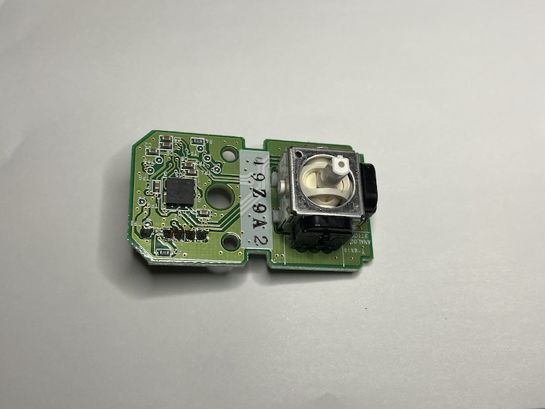 May include: A green circuit board with a small black chip and a silver and black analog stick. The board has the number "2 979A 2 1" printed on it.