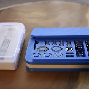 May include: Two cases on a wooden surface. One is a clear plastic case containing a syringe. The other is a blue case with a hinged lid, holding small metal and plastic components, including springs and rings.